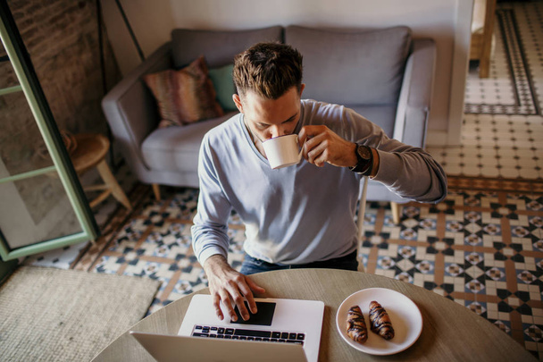 Handsome coworker man working at living room at home. Man sitting at wooden table using laptop and mobile phone. Blurred background. - Фото, зображення
