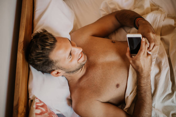 Carefree guy enjoying new day.Handsome young man in bed typing on cell phone, sending text message or dialing number - Fotografie, Obrázek
