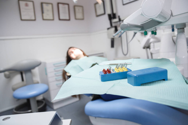 Молодая женщина, сидящая на стоматологическом стуле в комнате клиники в мягком фокусе с оборудованием и инструментами для очистки рта и для удаления полостей в фокусе
. - Фото, изображение