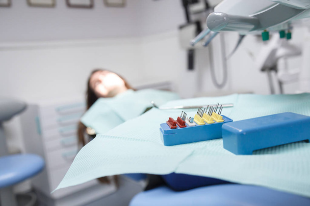 Jovem mulher sentada em cadeira dentária na sala de clínica em foco suave com equipamentos e instrumentos para limpeza da boca e para remoção de cavidades em foco
. - Foto, Imagem