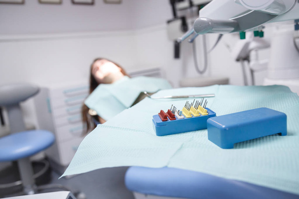 Молодая женщина, сидящая на стоматологическом стуле в комнате клиники в мягком фокусе с оборудованием и инструментами для очистки рта и для удаления полостей в фокусе
. - Фото, изображение