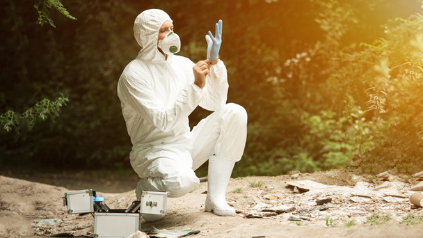 selective focus of male scientist in protective mask and suit putting on latex glove in forest - Фото, зображення