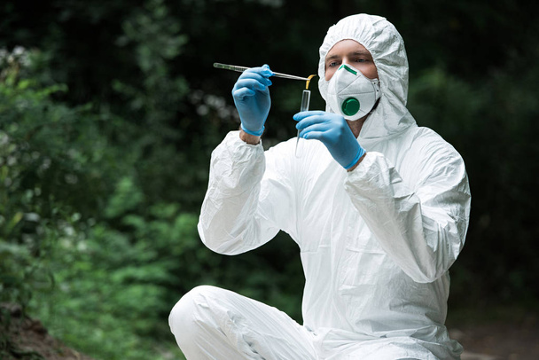 male scientist in protective mask and suit putting sample of yellow leaf by tweezers in test flask outdoors  - Photo, image
