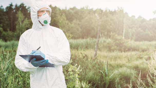 male scientist in protective mask, googles and suit looking away and writing in clipboard in meadow  - Valokuva, kuva