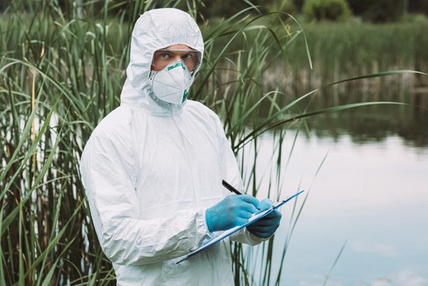 científico masculino en máscara protectora y escritura de traje en portapapeles cerca del agua al aire libre
  - Foto, Imagen