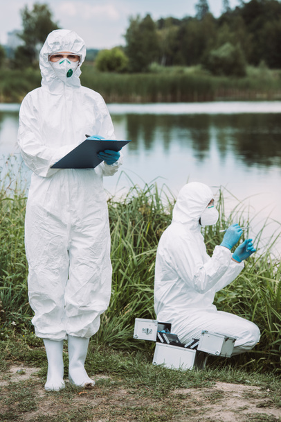 female scientist in protective mask and suit writing in clipboard while her colleague putting sample of water near river outdoors  - Photo, Image