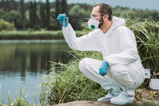 vista lateral del científico masculino en traje protector que examina la muestra de agua en el matraz de ensayo al aire libre
  - Foto, Imagen
