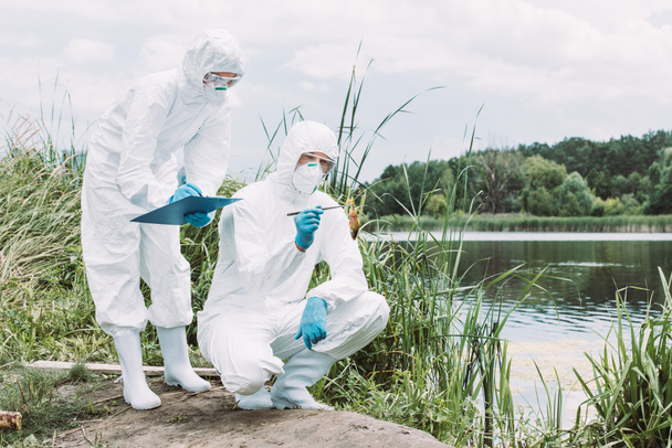 two scientists in protective masks and suits examining fish and writing in clipboard near river  - Foto, Bild