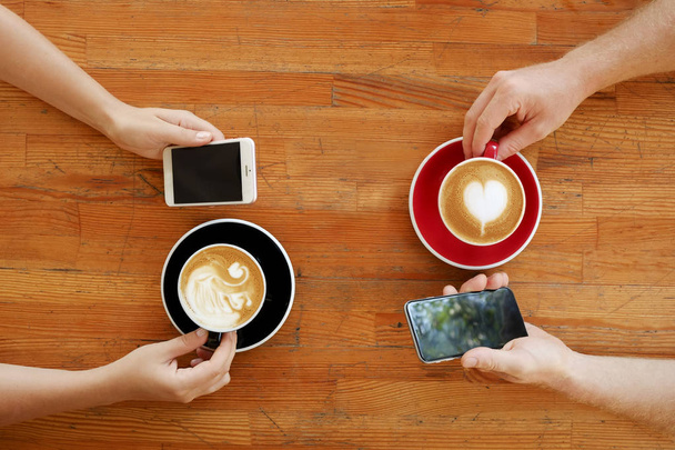Young couple on boring date at coffee shop constantly checking phone. Man & woman drinking cappuccino from different black & red cups holding blank screen smartphones. Background, top view, copy space - Foto, immagini