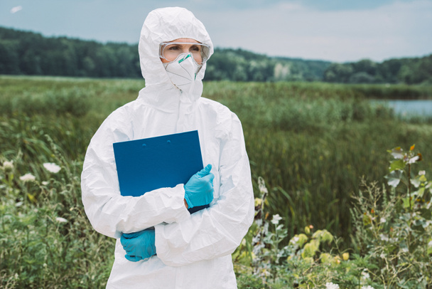 selective focus of female scientist in protective suit and googles holding clipboard in meadow  - Photo, Image