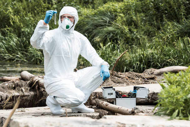 científico masculino en máscara protectora y traje que examina la muestra de agua en el frasco de prueba al aire libre
  - Foto, Imagen