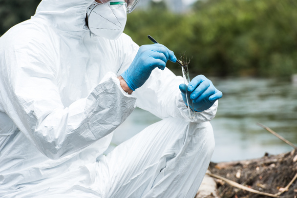cropped image of male scientist in protective suit and mask putting sample of soil by tweezers in test flask outdoors  - 写真・画像