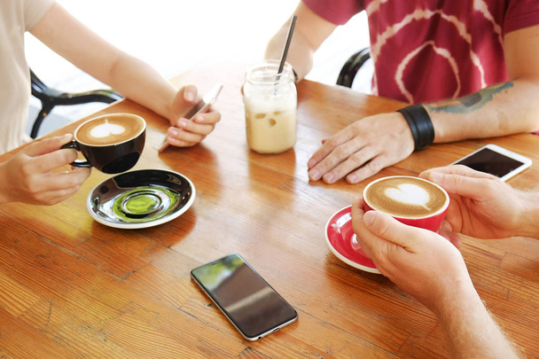 Group of friends drinking cappuccino at coffee shop, staring at smartphones, checking likes & messages. Addiction to social media. Young men & women holding cell phones & drinks. Background, top view. - Photo, Image
