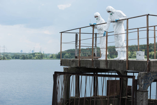 male scientist in protective suit and mask pointing at water to female colleague with laptop outdoors  - Fotoğraf, Görsel