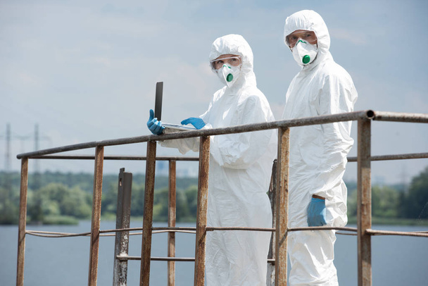 two scientists in protective suits and masks working with laptop and looking at camera near river outdoors  - Fotó, kép
