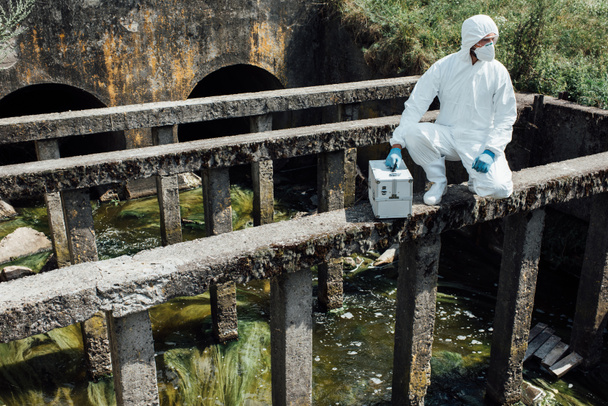 male scientist in protective mask and suit sitting with working suitcase near sewerage  - Photo, Image