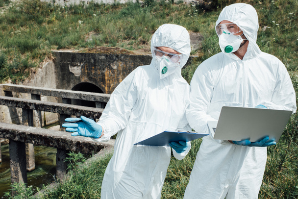 selective focus of female scientist with clipboard pointing to male colleague with laptop near sewerage  - Photo, Image