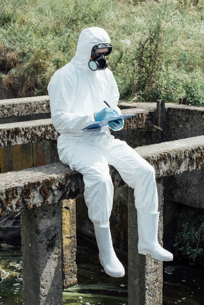 selective focus of male scientist in protective mask and suit writing in clipboard while sitting near sewerage  - Foto, imagen