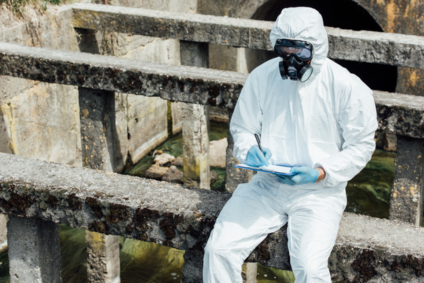  male scientist in protective mask and suit writing in clipboard while sitting near sewerage  - Photo, Image