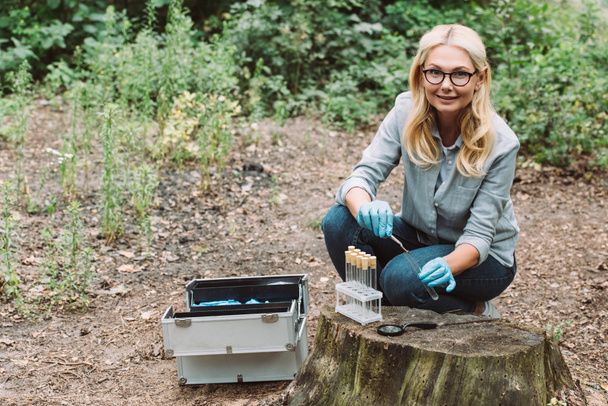 smiling female scientist putting twig by tweezers in test flask in forest  - Photo, Image