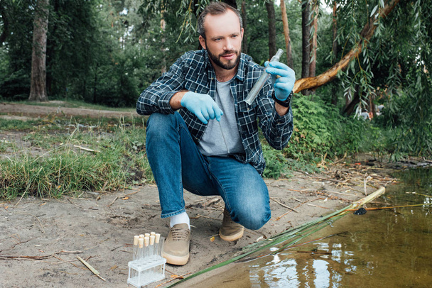 ученый-мужчина в латексных перчатках берет образец воды в тестовой фляжке на открытом воздухе
  - Фото, изображение