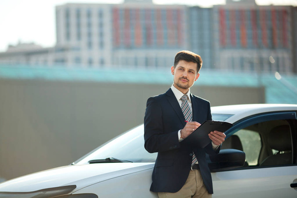 handsome young man in a suit standing near a white car on the street signing documents - Fotografie, Obrázek