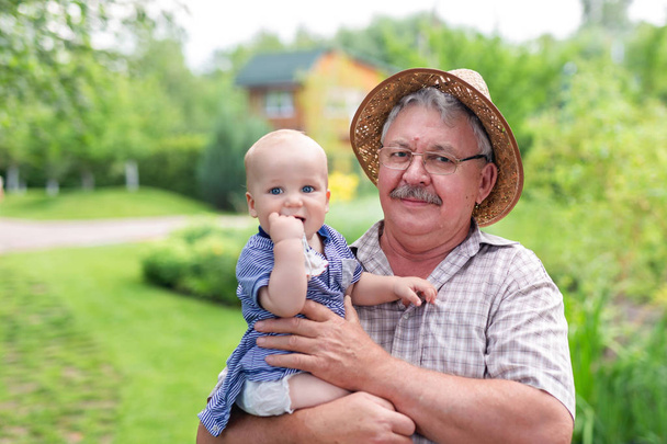 Senior gray man in straw hat holding grandson on hand and having fun in green summer garden. Baby boy with grandgather enjoying together outdoors rural cottage backyard.Generations relations concept - Photo, Image