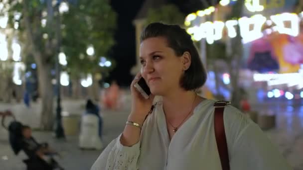 A steadicam shot of a pretty young woman talking on the phone and smiling at night with a glowing amusement park on the background - Кадры, видео