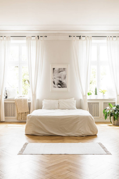 Poster above white bed in spacious bright bedroom interior with windows and rug. Real photo - Foto, Imagen