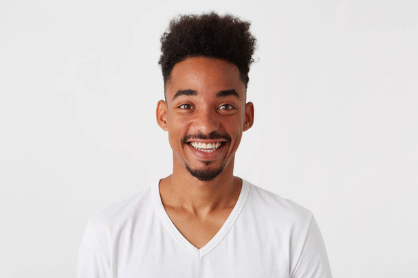 Портрет веселого привлекательного африканского молодого человека с афропрической носит футболку выглядит счастливым и улыбающимся изолированным на белом фоне
 - Фото, изображение