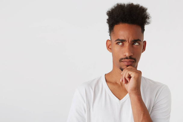 Portret van doordachte knappe Afro-Amerikaanse jonge man met afro kapsel draagt t shirt ziet er nadenkend en denken geïsoleerd op witte achtergrond - Foto, afbeelding