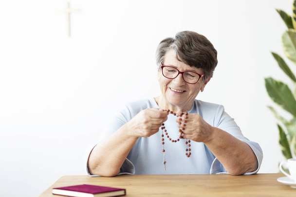 Smiling senior woman holding rosary while sitting at table with bible - Fotoğraf, Görsel
