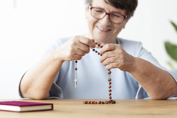 Smiling religious elderly woman praying with rosary at table with bible. Focus on hands - Foto, afbeelding