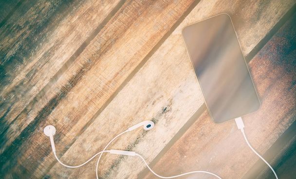 A top view of a set of wired earbud headphones and mobile phone lying on a rustic wooden table. Styling and grain effect added to image. - Photo, Image
