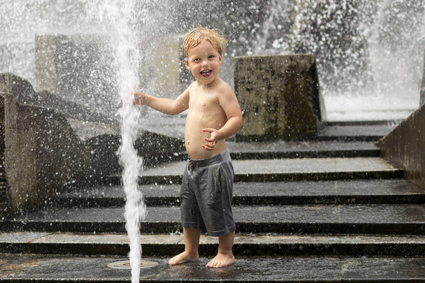 Grappige babyjongen probeert te cauch water stroom in de fontein. Schattig peuter spelen in de stad-fontein. - Foto, afbeelding