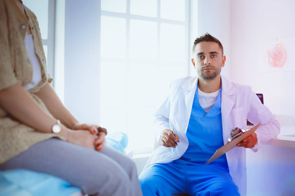 Bello medico sta parlando con giovane paziente femminile e prendere appunti mentre seduto nel suo ufficio - Foto, immagini