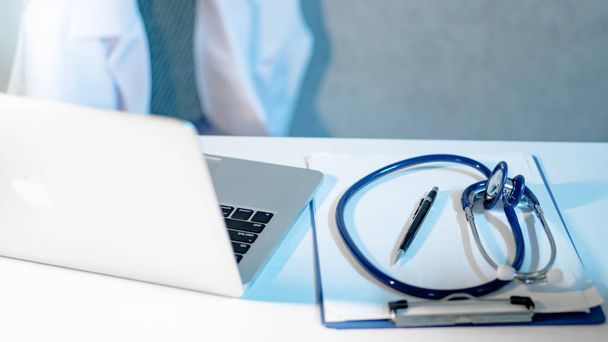 Blue stethoscope, clipboard, pen and laptop computer on doctor desk. Blurred white doctor coat in the background. Medical treatment equipment and health care diagnostic concepts - Photo, Image