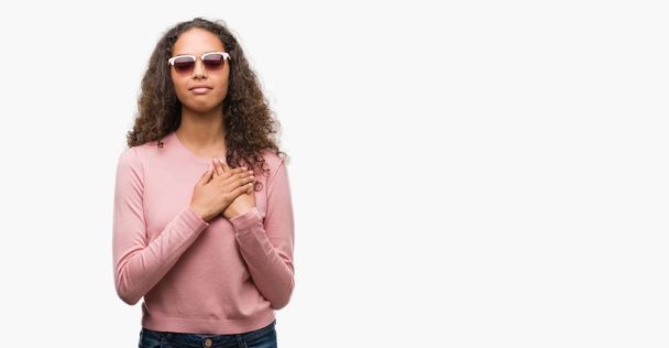 Beautiful young hispanic woman wearing sunglasses smiling with hands on chest with closed eyes and grateful gesture on face. Health concept. - Foto, immagini