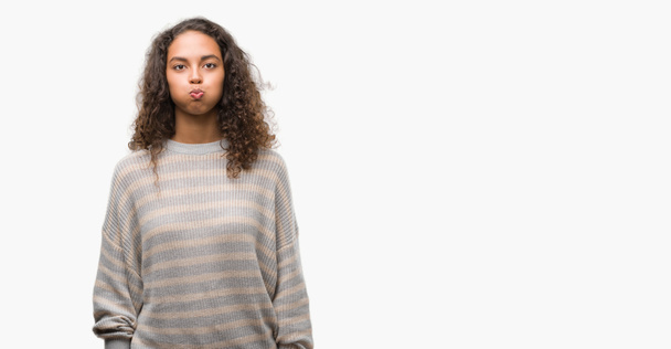 Beautiful young hispanic woman wearing stripes sweater puffing cheeks with funny face. Mouth inflated with air, crazy expression. - Photo, Image