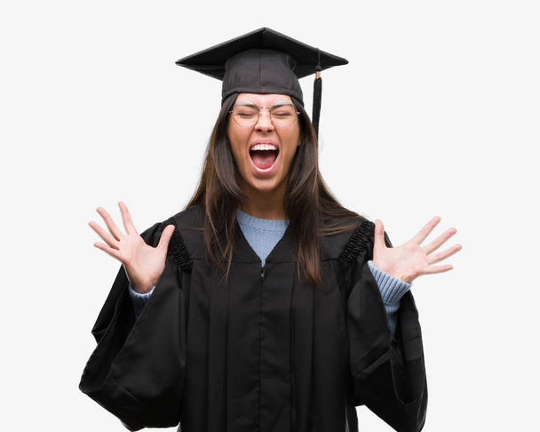 Young hispanic woman wearing graduated cap and uniform celebrating mad and crazy for success with arms raised and closed eyes screaming excited. Winner concept - Foto, afbeelding