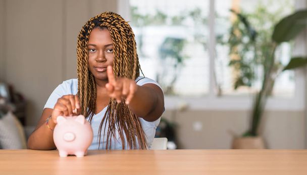 Afrikkalainen amerikkalainen nainen säästää rahaa säästöpossu pankki osoittaa sormella kameraan ja sinulle, käsi merkki, positiivinen ja luottavainen ele edestä
 - Valokuva, kuva
