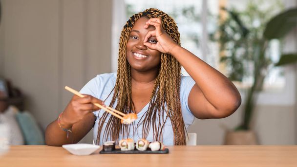 Mujer afroamericana comiendo sushi con la cara feliz sonriendo haciendo bien signo con la mano en el ojo mirando a través de los dedos
 - Foto, imagen