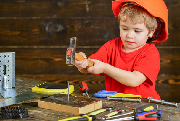 Handcrafting concept. Toddler on busy face plays with hammer tool at home in workshop. Kid boy play as handyman. Child in helmet cute playing as builder or repairer, repairing or handcrafting - Foto, imagen