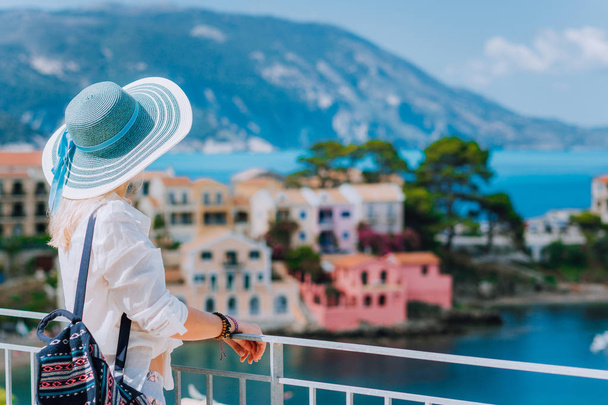 Tourist woman wearing blue sunhat and white clothes enjoying view of colorful tranquil village Assos on sunny day. Stylish female visiting Kefalonia in summer time on Greece travel vacation - Фото, зображення