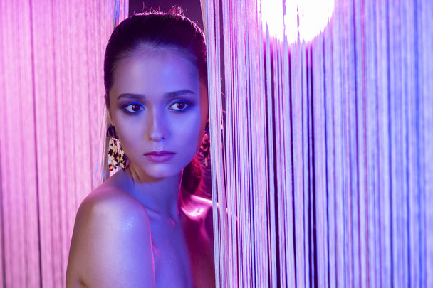 A beautiful glamorous brunette girl with naked shoulders wearing large elegant earrings peeps through a translucent curtain made of white threads. Multicolored artistic stage lighting. Copy space. - Foto, imagen
