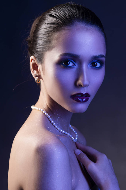 Beautiful brunette girl with naked shoulders and dark red lips make-up, wearing earrings and a pearl necklace, illuminated in blue. Conceptual low key photo. Copy space. Advertising design of jewelry. - Foto, Bild