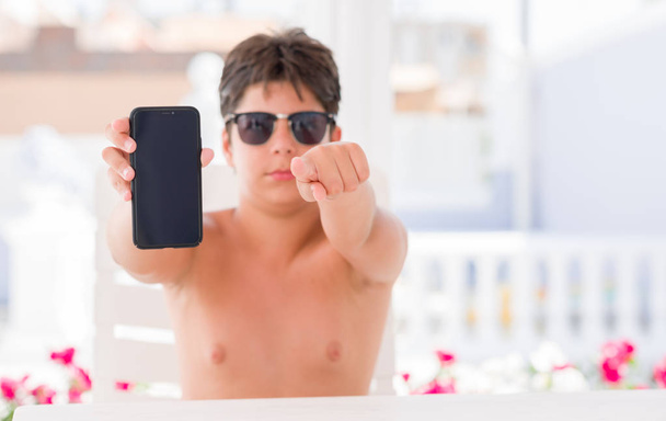 Kleinkind im Urlaub zeigt Smartphone mit Zeigefinger auf Kamera und Sie, Handzeichen, positive und selbstbewusste Geste von vorne - Foto, Bild