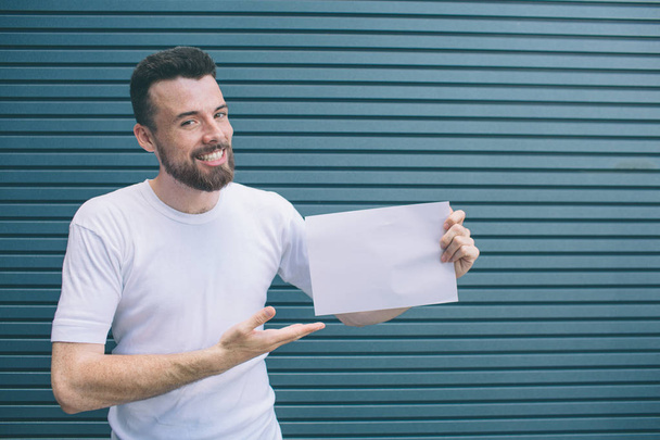 Nice and positive man iss standing and smiling. He is posing and looking at camera. Guy is holding piece of paper and poiting on it with hand. Isolated on striped and blue background. - Foto, Imagen