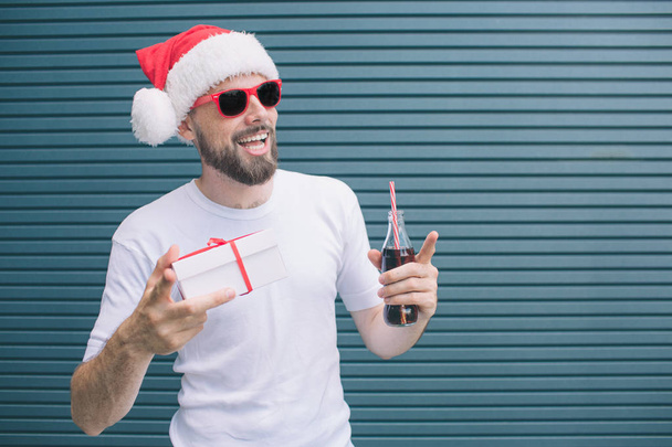 Nice man is standing and holding present in one hand and bottle of coke in another one. Also guy is pointing with finger. Man wears glasses and hat. Isolated on striped and blue background. - Zdjęcie, obraz