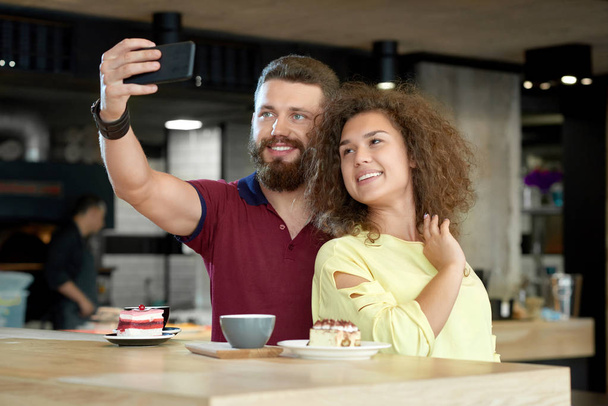Widok z przodu uśmiechnięta para biorąc selfie podczas dnia w kawiarni. - Zdjęcie, obraz